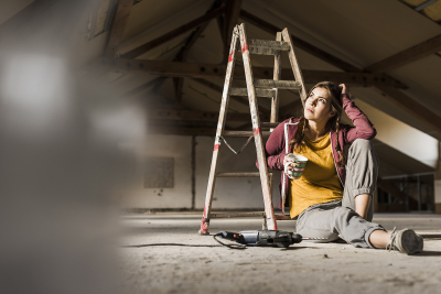 Frau vor Leiter mit Tasse in der Hand auf renovierungsbedürftigem Dachboden.