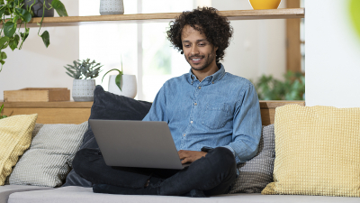 Mann sitzt lächelnd auf Bank im Schneidersitz mit Laptop. Die digitale Aufwertung von Immobilien mit Möbeln und Deko-Elementen ist der Kern des Virtual Stagings.