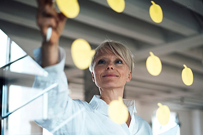 Frau klebt zuversichtlich schauend Postits an eine gläserne Wand Frau klebt zuversichtlich schauend Postits an eine gläserne Wand